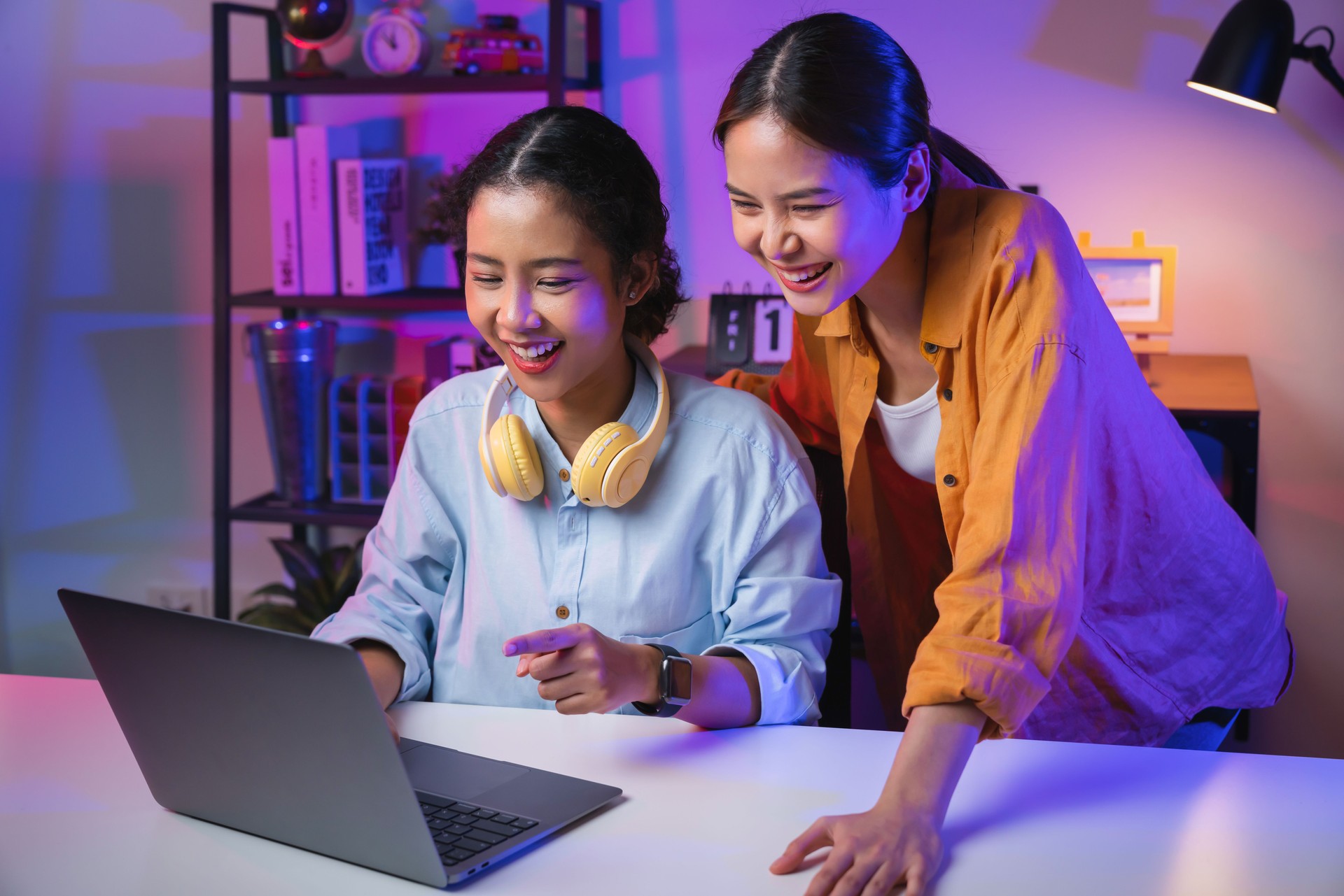 Two happy female students in casual wear wireless headphones using social media on internet with laptop at home.